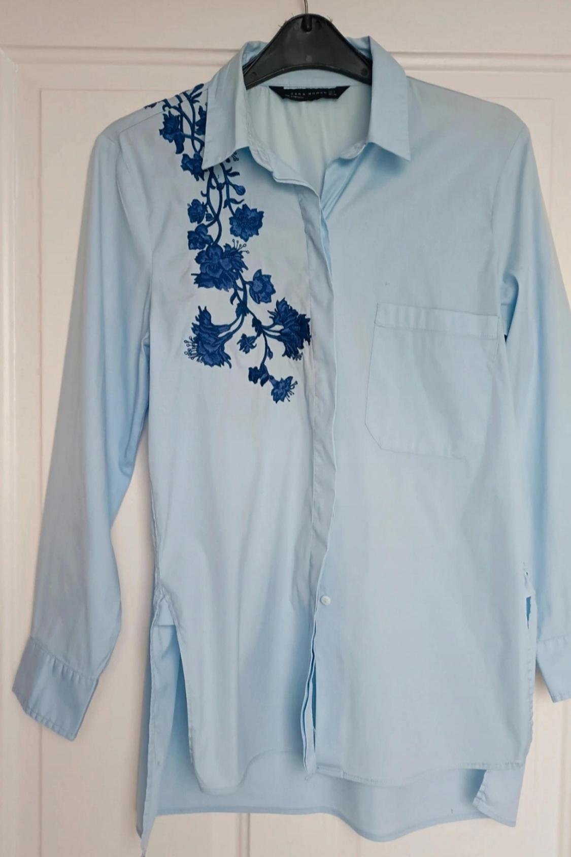 Front view of a light blue shirt with blue floral embroidery on the left shoulder and a front pocket.