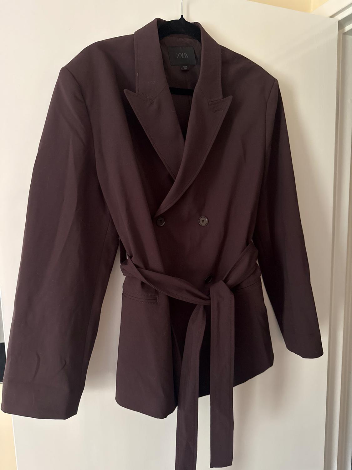 Dark brown double-breasted blazer with a tied belt, hanging on a black hanger over a white door. Label with the text ZARA.