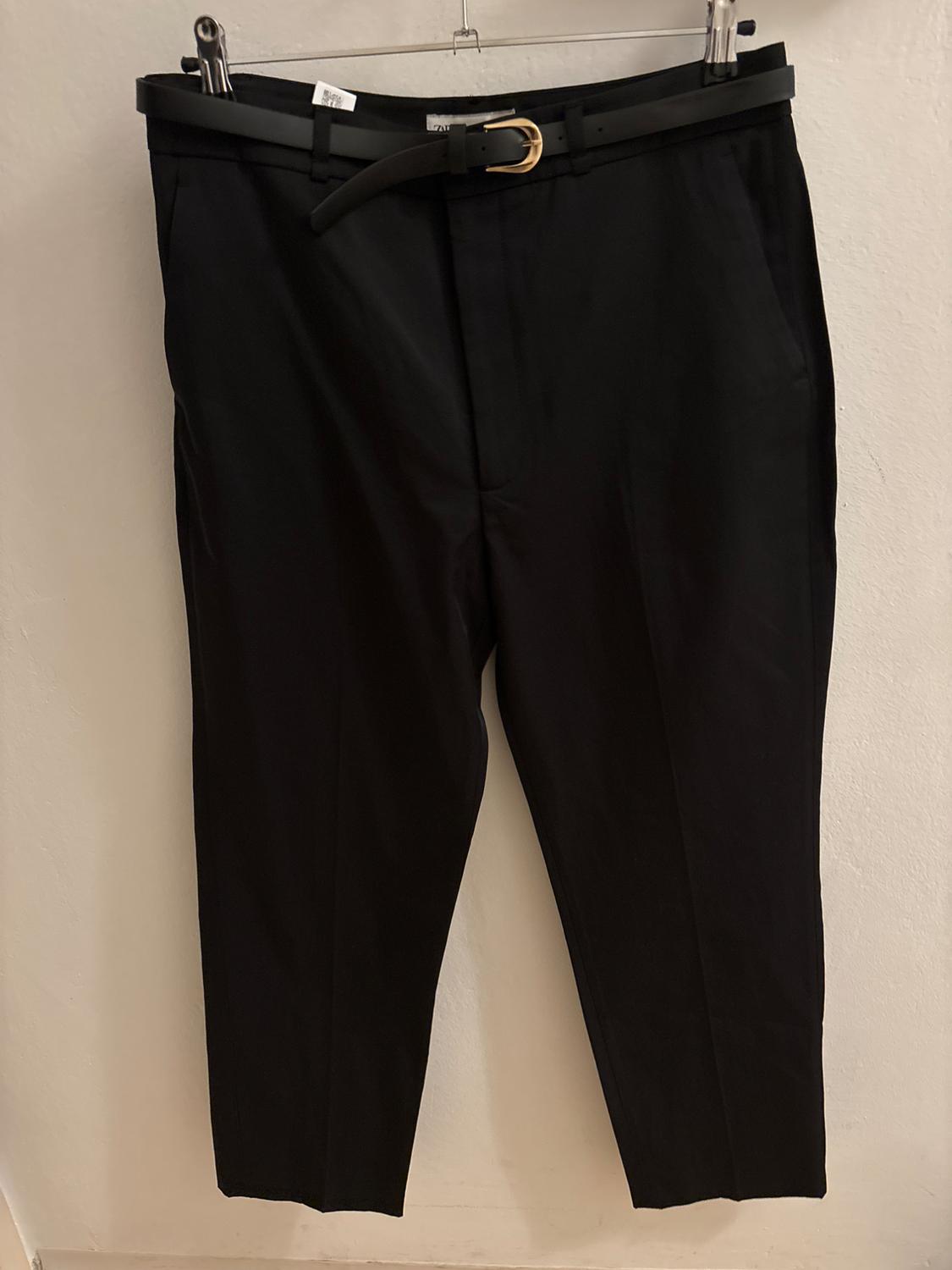 Black trousers hanging on a hanger with a black belt and gold buckle. Plain white background.