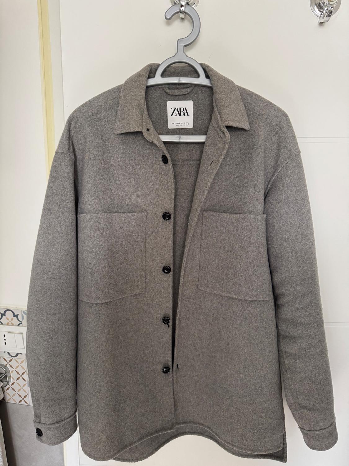 Grey wool jacket hanging on a hanger, with black buttons and two front pockets. Visible label with the text "ZARA".