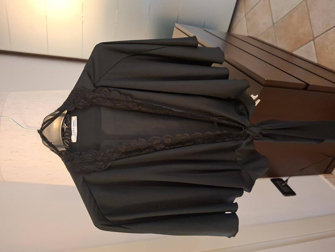 Black lace jacket hanging on a hanger, against a white wall and wooden furniture background. Visible tag: "Zara Woman".