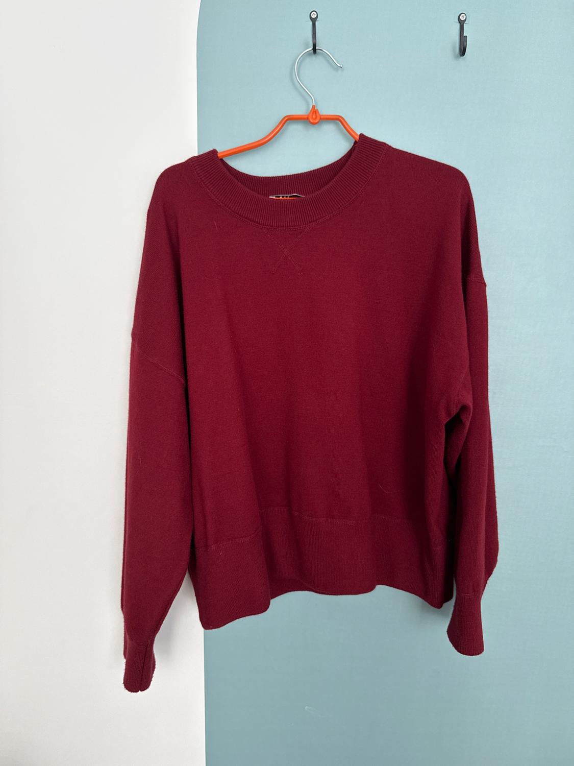 Burgundy crew neck jumper on an orange hanger, hanging on a wall divided into white and light blue. Casual and minimalist style.