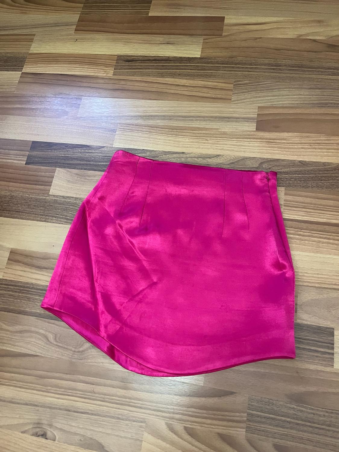 Fuchsia satin mini skirt with an asymmetric hem, darts and a side zip, laid out on a wooden floor. Casual style.