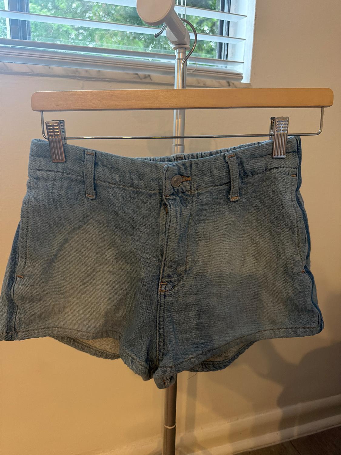 Light blue denim shorts hanging on a wooden hanger with clips in front of a window with white blinds.