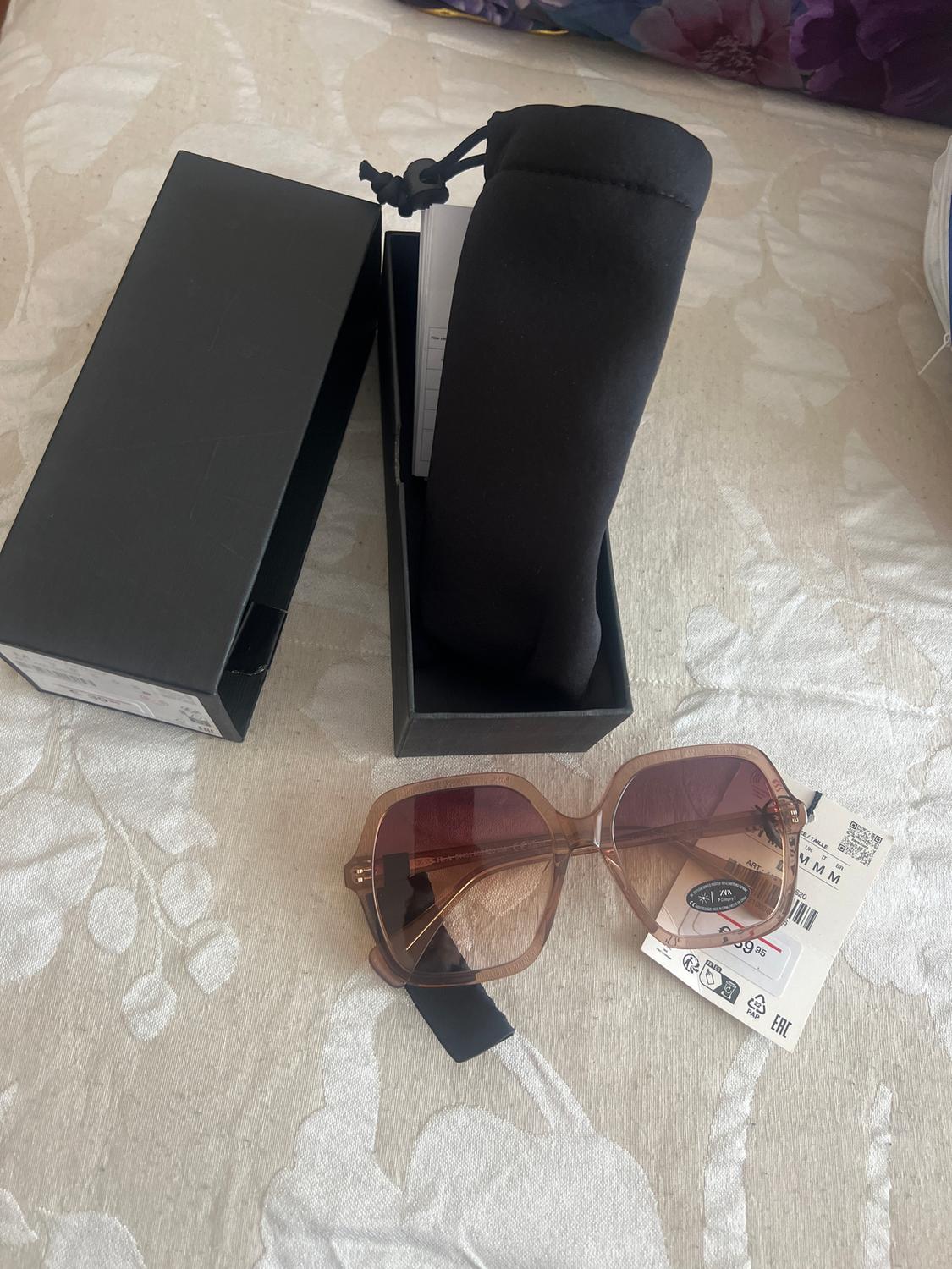 Brown-framed sunglasses with tinted lenses on white fabric. Black box and black fabric pouch alongside. Visible label.