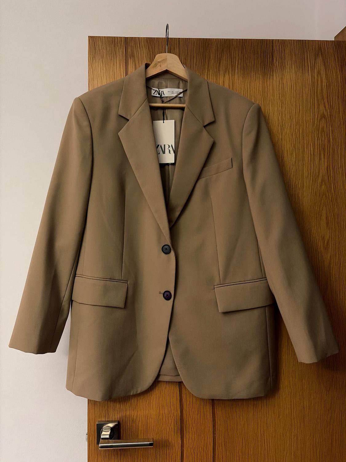 Brown jacket hanging on a wooden door. Visible label with the text "ZARA". Simple and elegant interior setting.