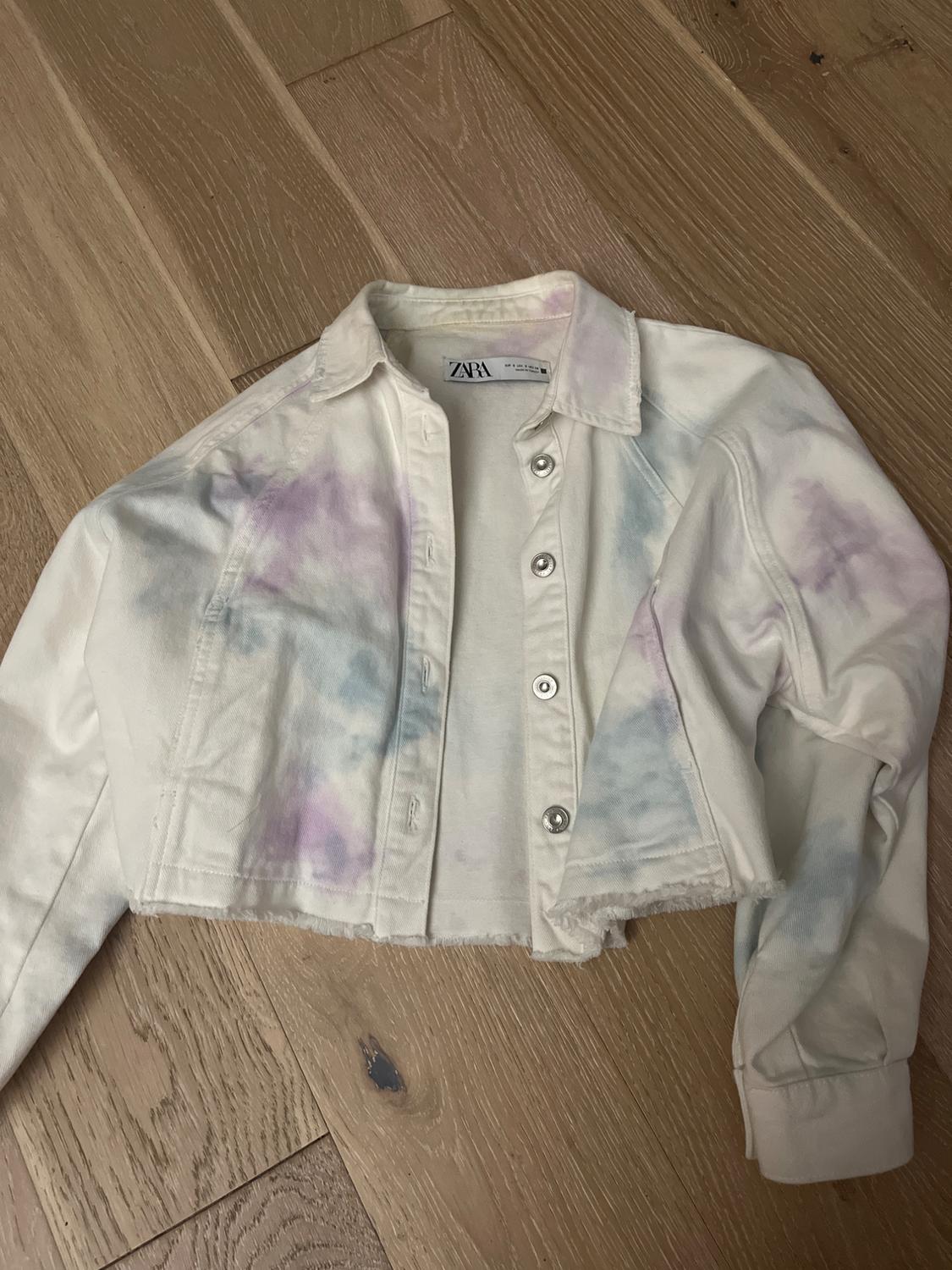 Short denim jacket with a tie-dye effect in pastel shades on a wooden floor. Visible label with "ZARA" text.
