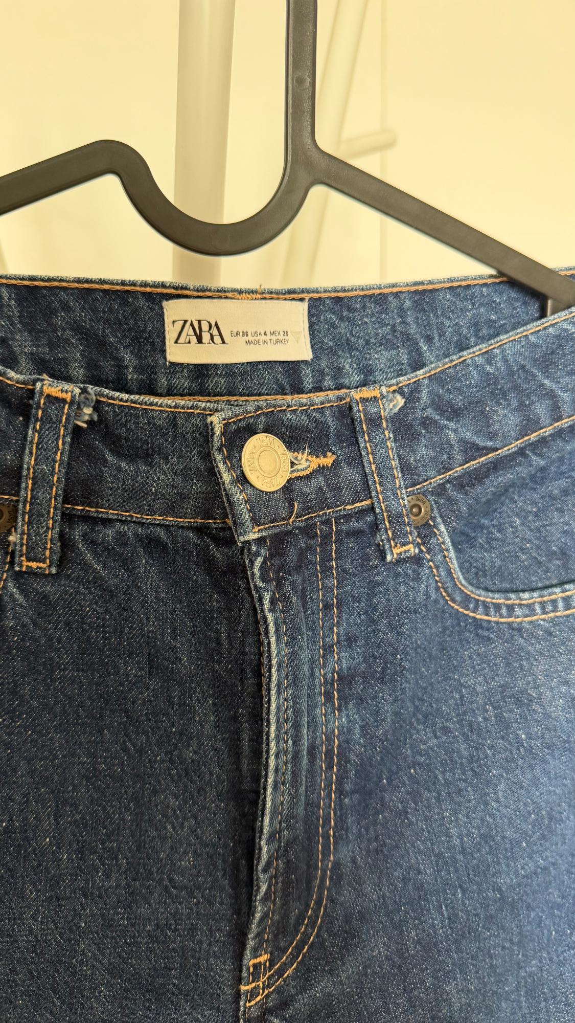 Detail of blue jeans on a black hanger. Label with the text ZARA EUR 36 USA 4 MEX 26 MADE IN TURKEY. Orange stitching and metallic button.