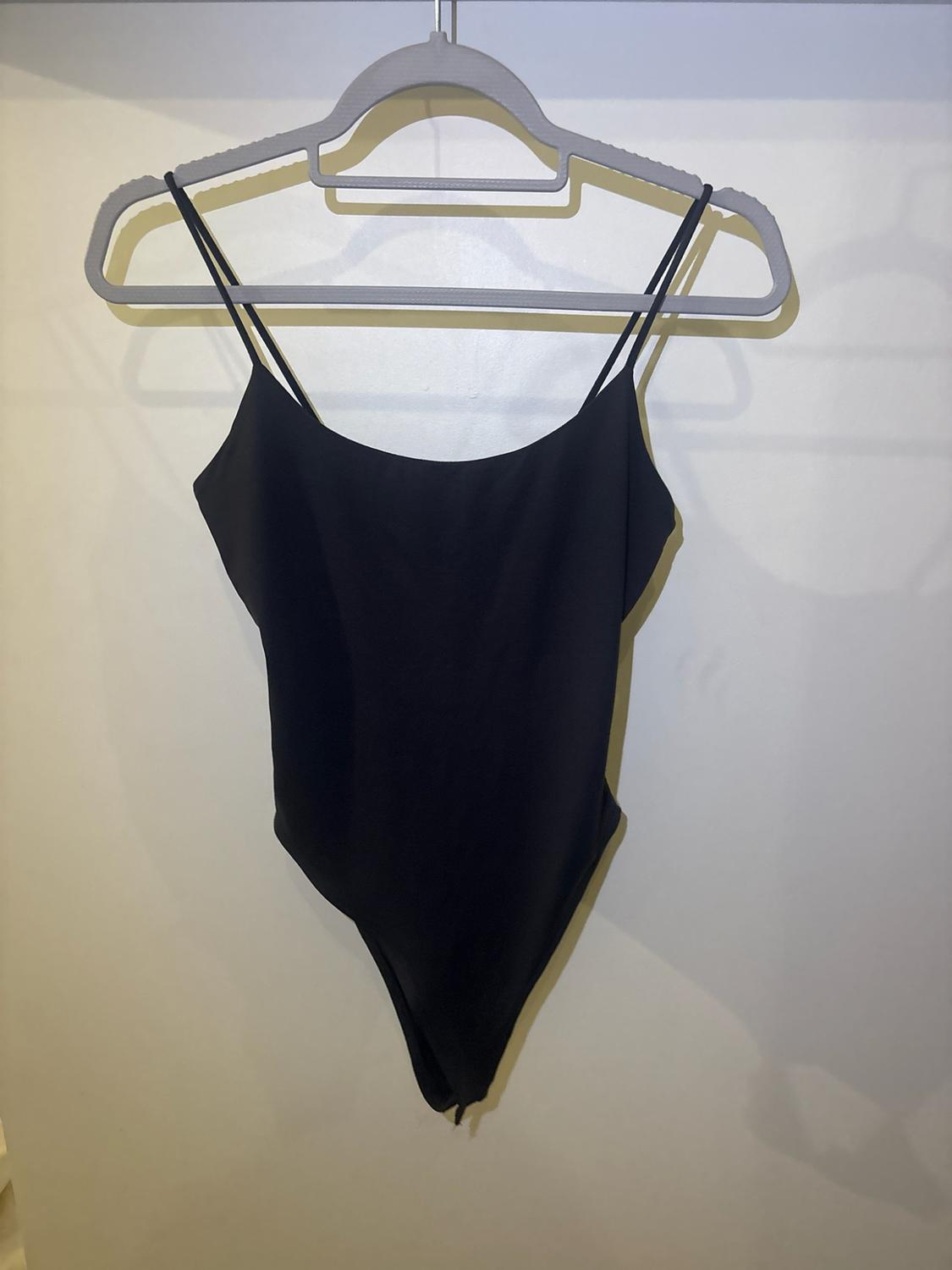 Plain black bodysuit with double spaghetti straps and a round neckline, hanging on a grey hanger against a light wall with shadows.