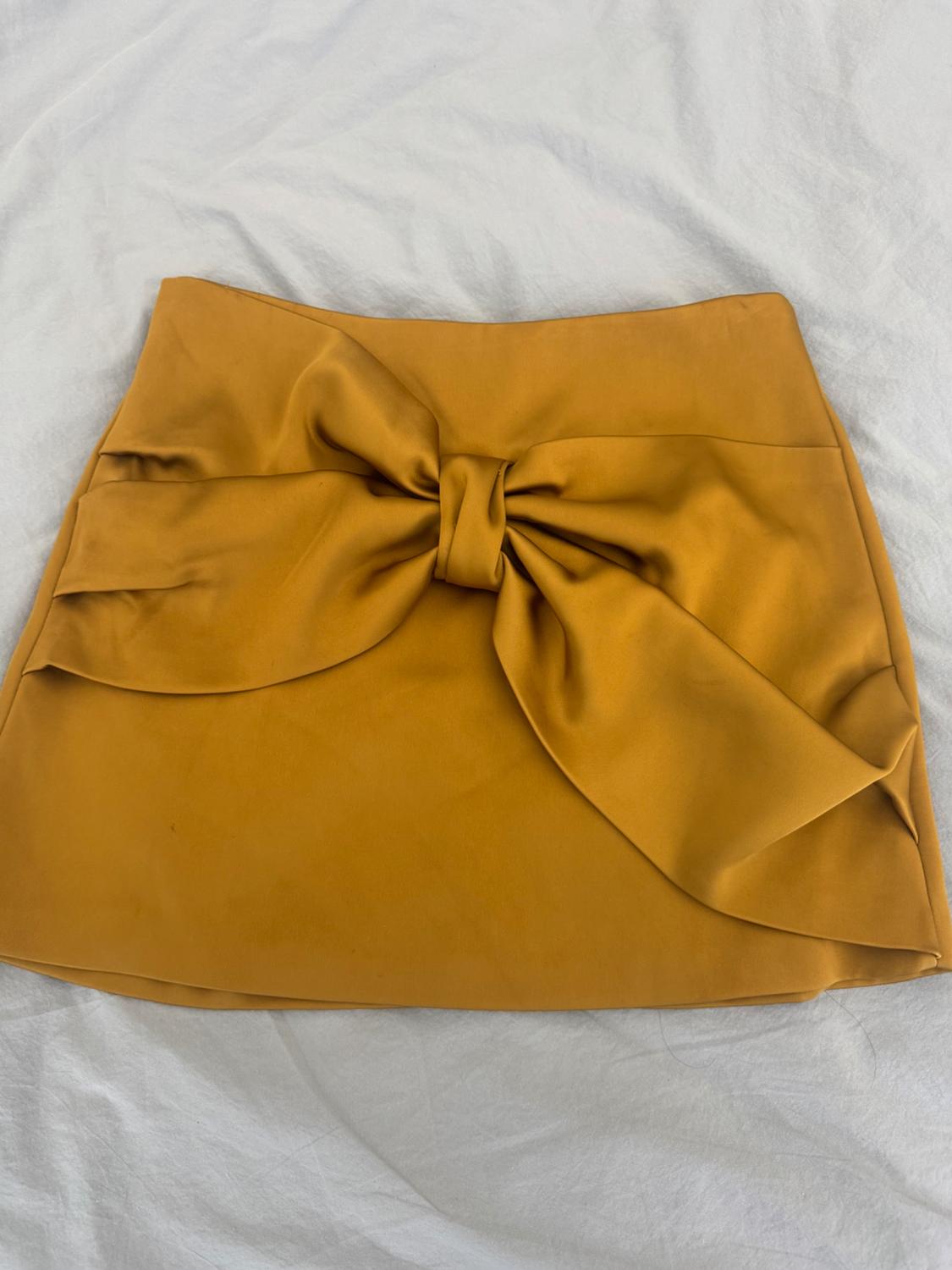 Satin mustard-coloured miniskirt with a large bow at the front, spread out on a crumpled white fabric.