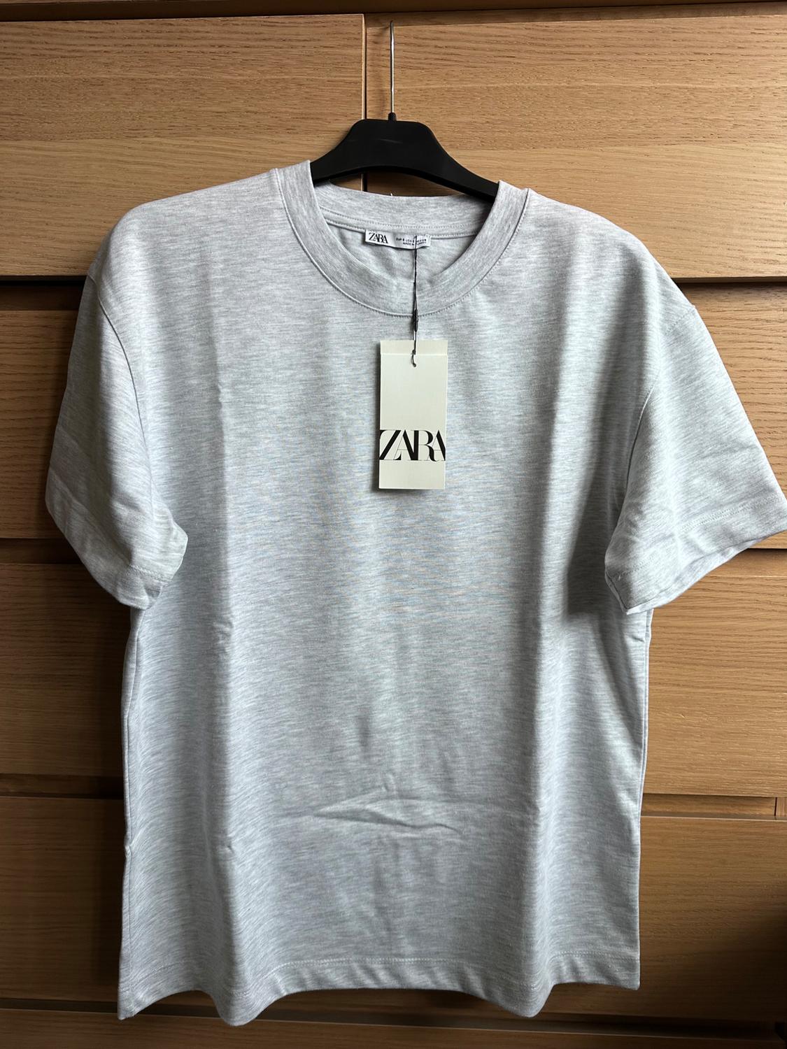 Light grey T-shirt hanging on a black hanger with a "ZARA" label, against a light wooden background.