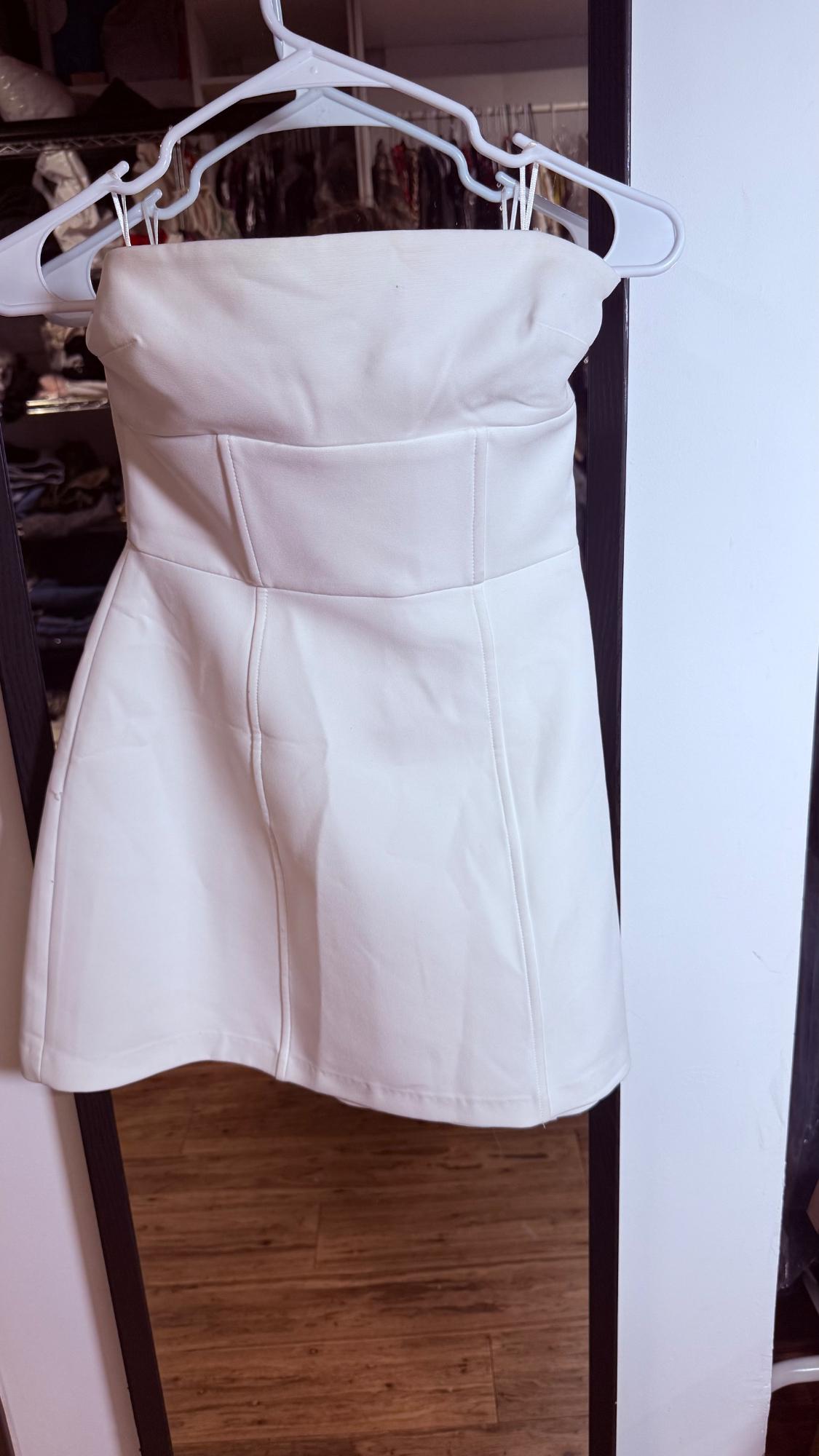 Short white strapless dress with exposed seams, hanging on a white hanger in front of a mirror reflecting a closet.