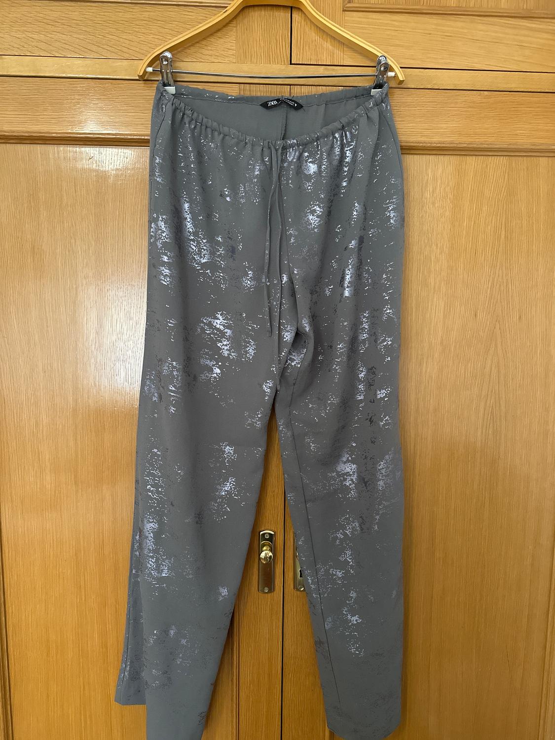 Grey trousers with a shiny print hanging on a wooden hanger in front of a light wood wardrobe door.