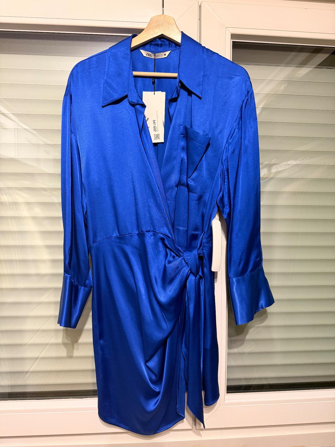Bright blue blouse with a tie at the waist and three-quarter length sleeves, hanging on a hanger in front of a window with blinds.