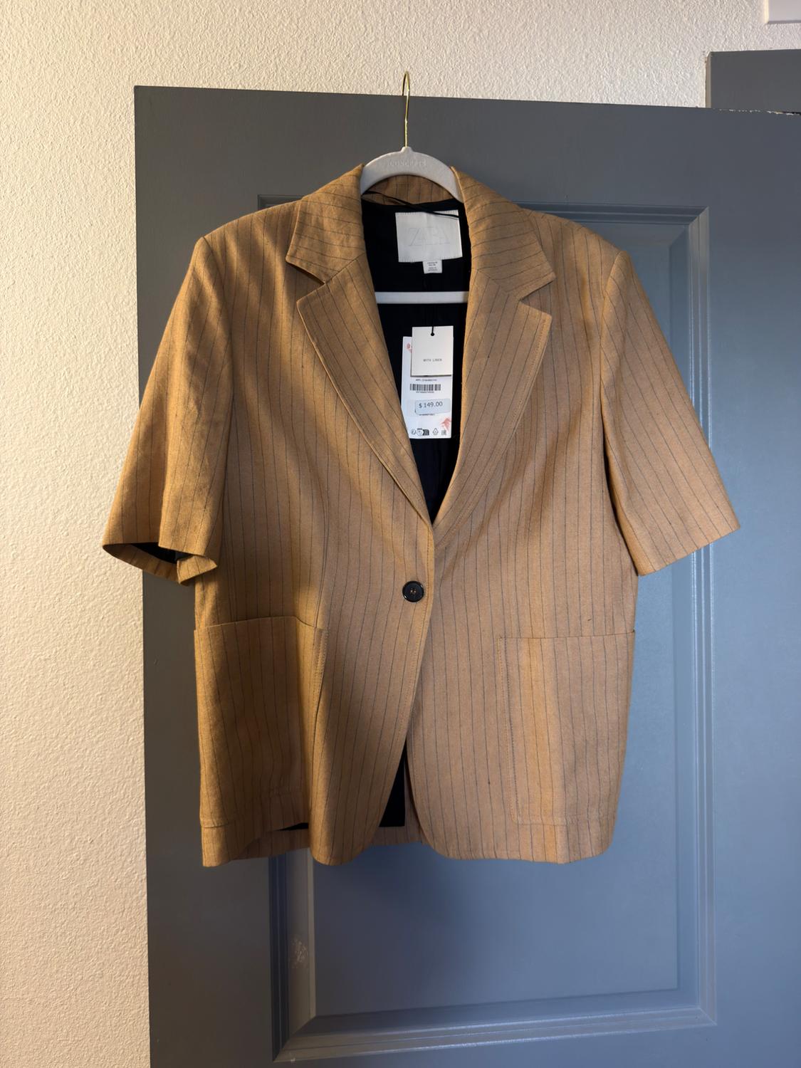 Camel short-sleeved blazer with fine stripes, hanging on a grey door. Labels with text ZARA, WITH LINEN and $149.00.