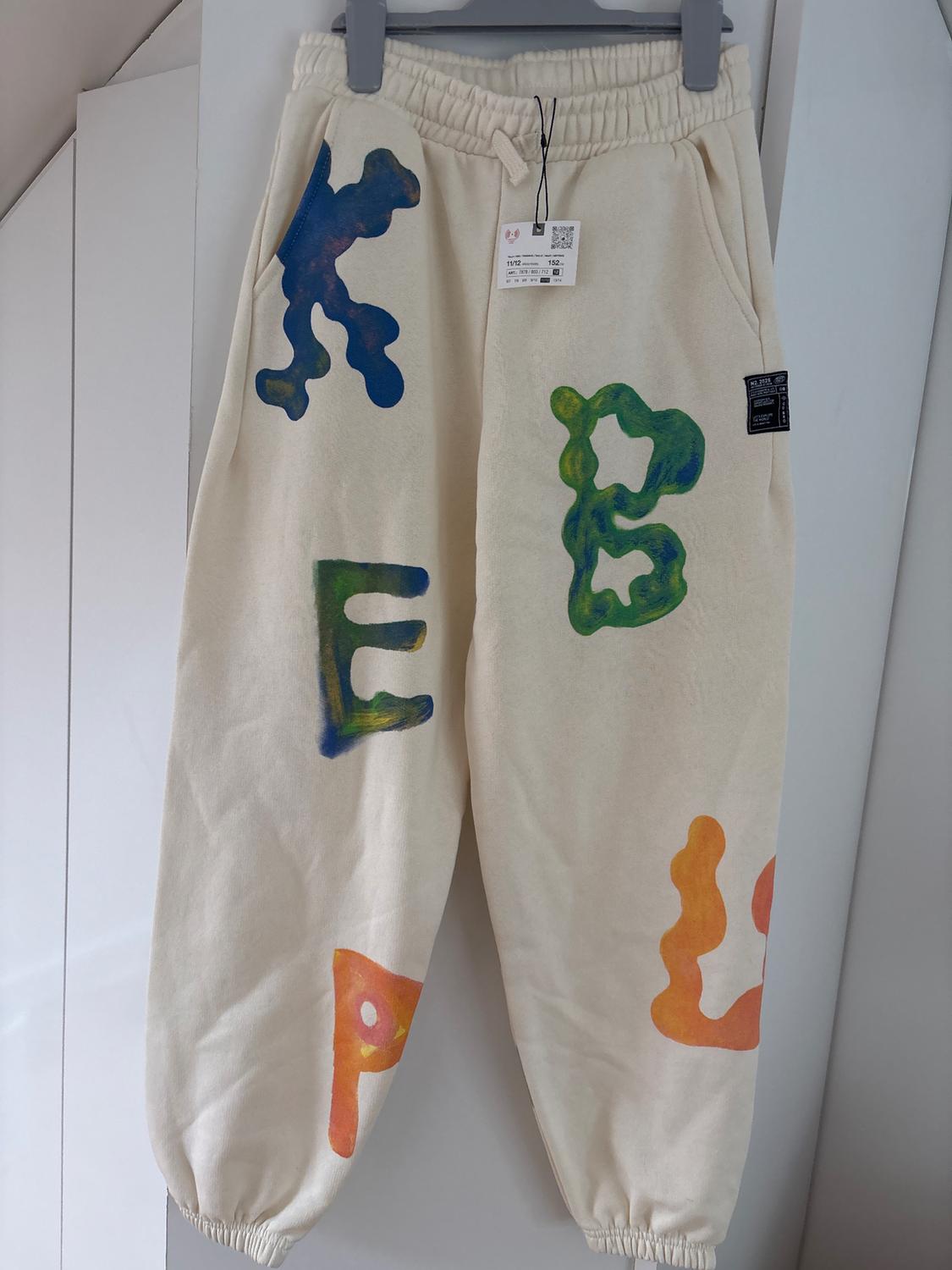 Beige trousers with colourful "KEEP" lettering in blue, green, and orange. Elasticated waist, hanging on a white hanger.