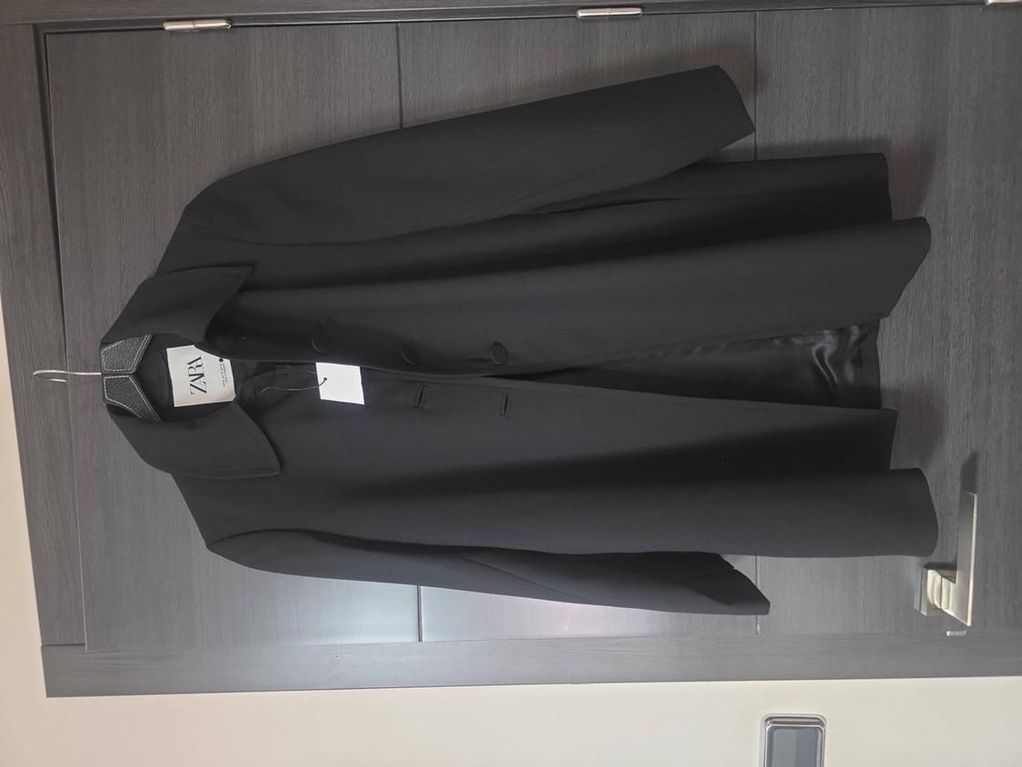 Black coat hanging on a grey wooden door. White label visible with the text ZARA. Relaxed and casual style.