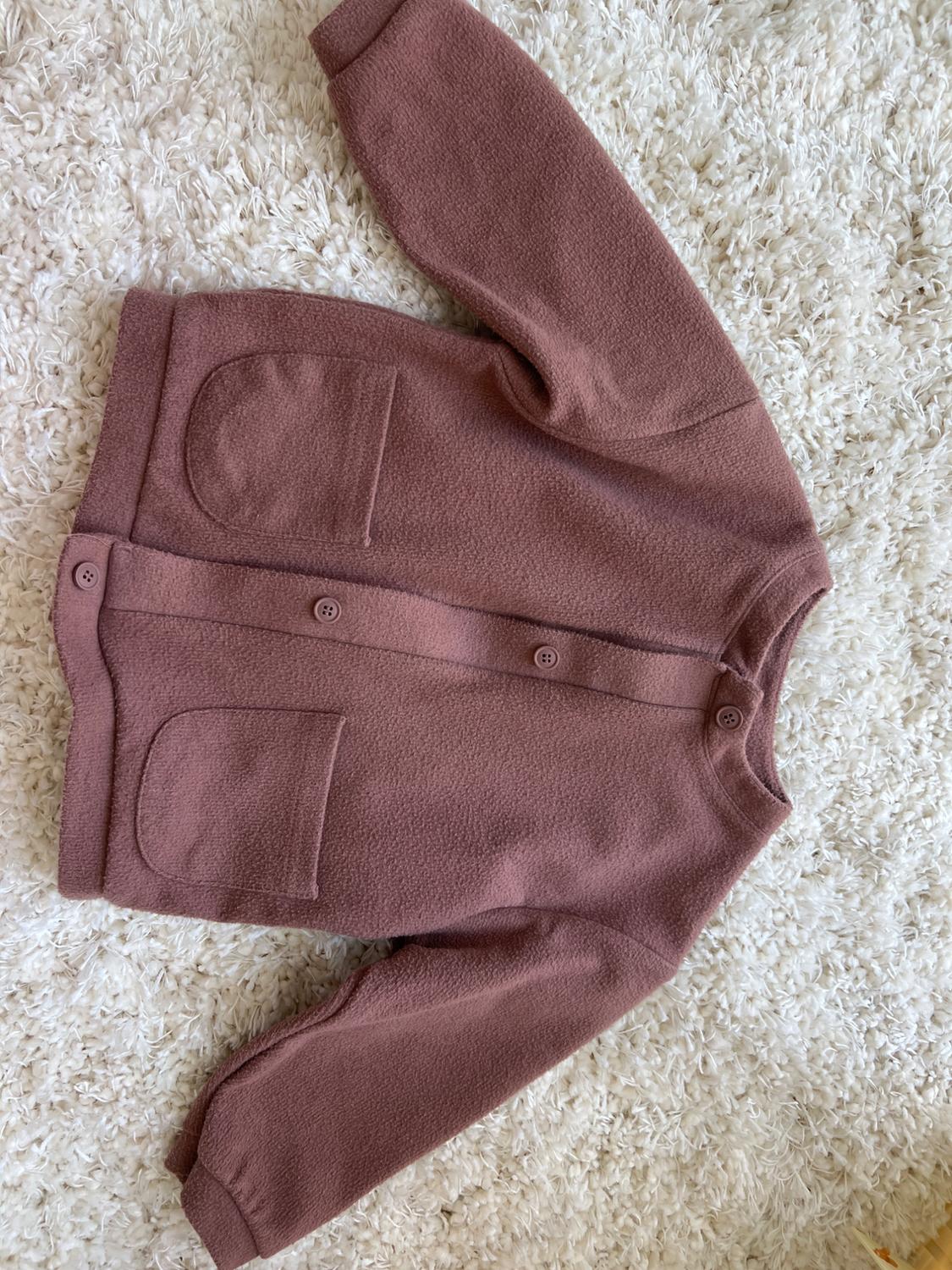 Pink wool jacket with buttons and two front pockets, placed on a soft textured white rug.