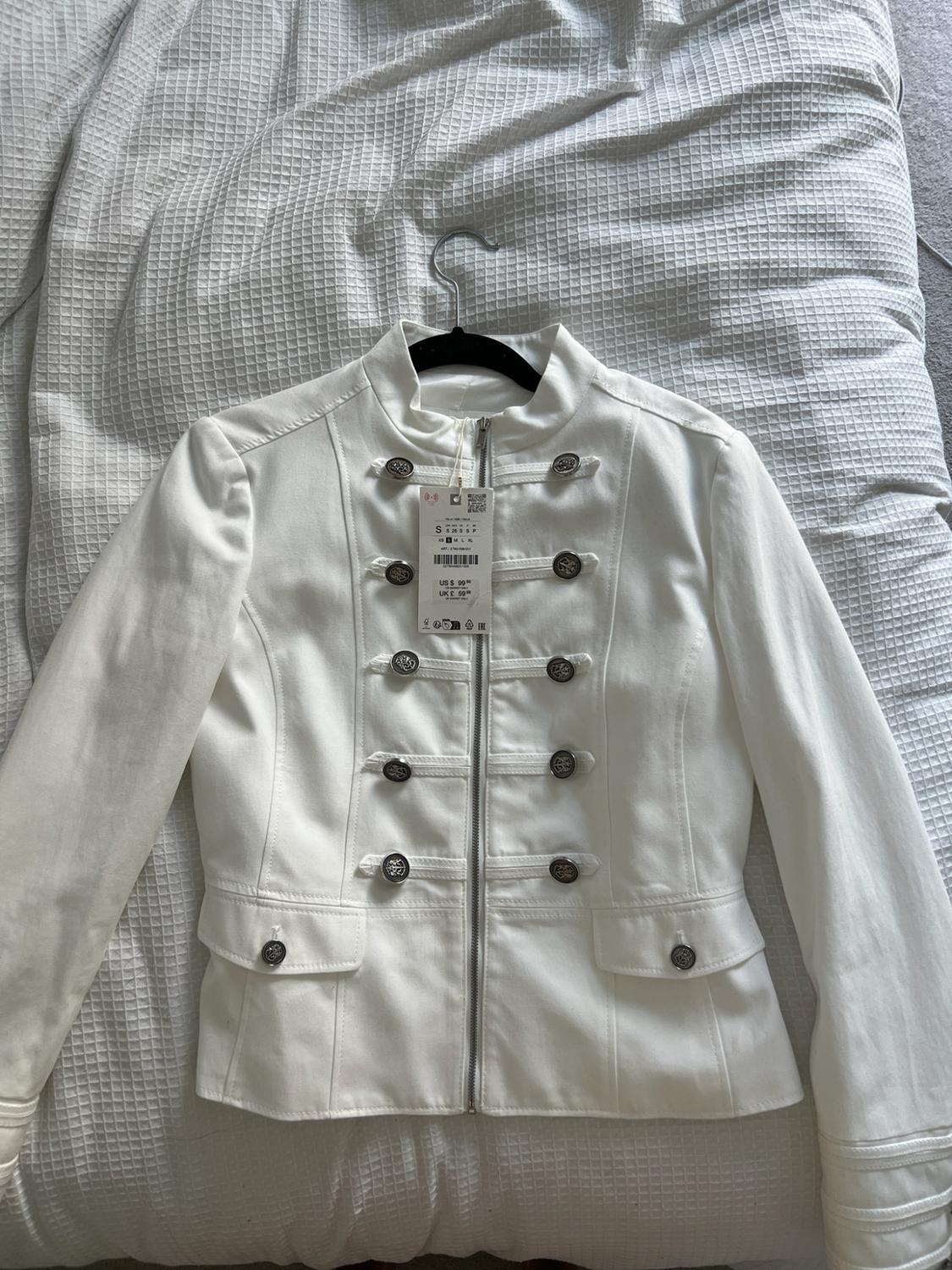 White military-style zipped jacket with buttons on a bed. Label with text US $ 99 UK £ 59.