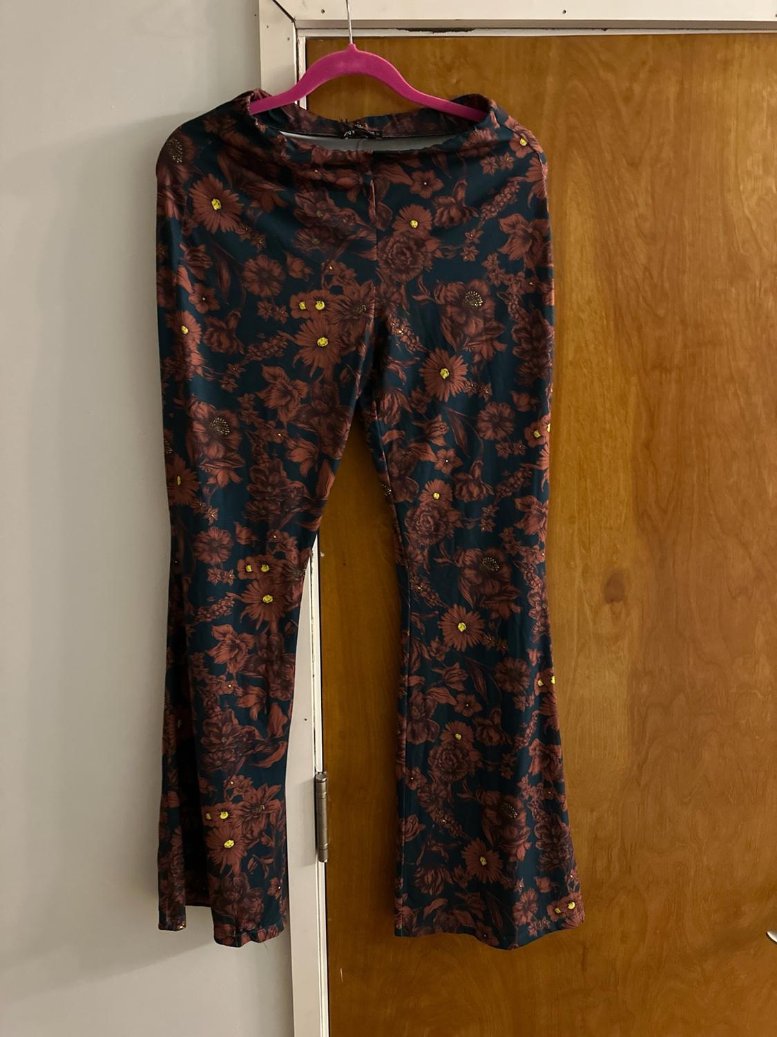 Dark flared trousers with a floral print and yellow shimmer, hanging on a pink hanger next to a wooden door.