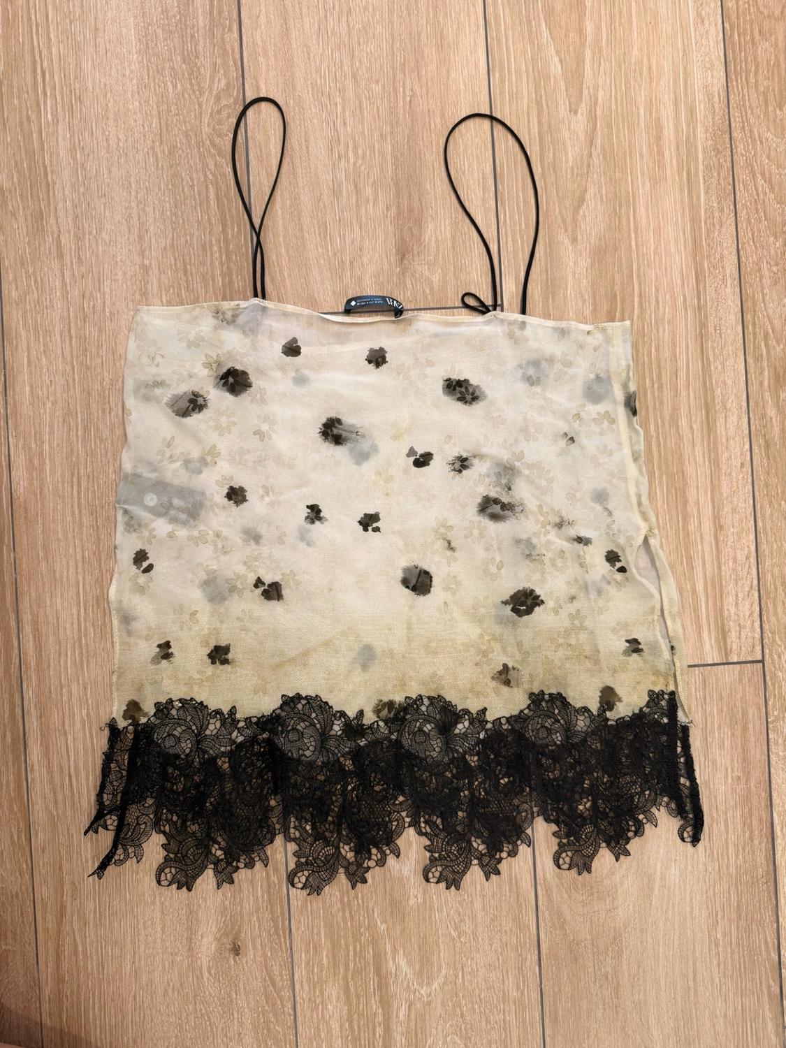 Beige semi-sheer top with dark print, thin straps and a black lace hem, laid out on a wooden floor.