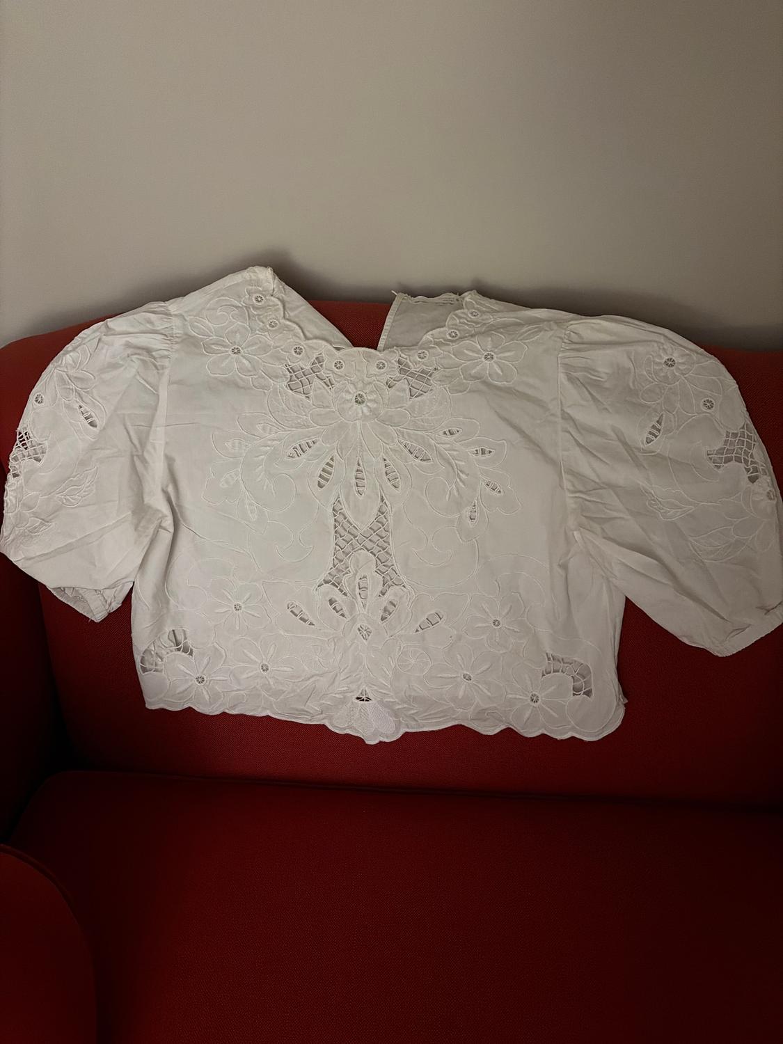 White blouse embroidered with flowers and lace, placed on a red sofa. Light gray wall in the background.