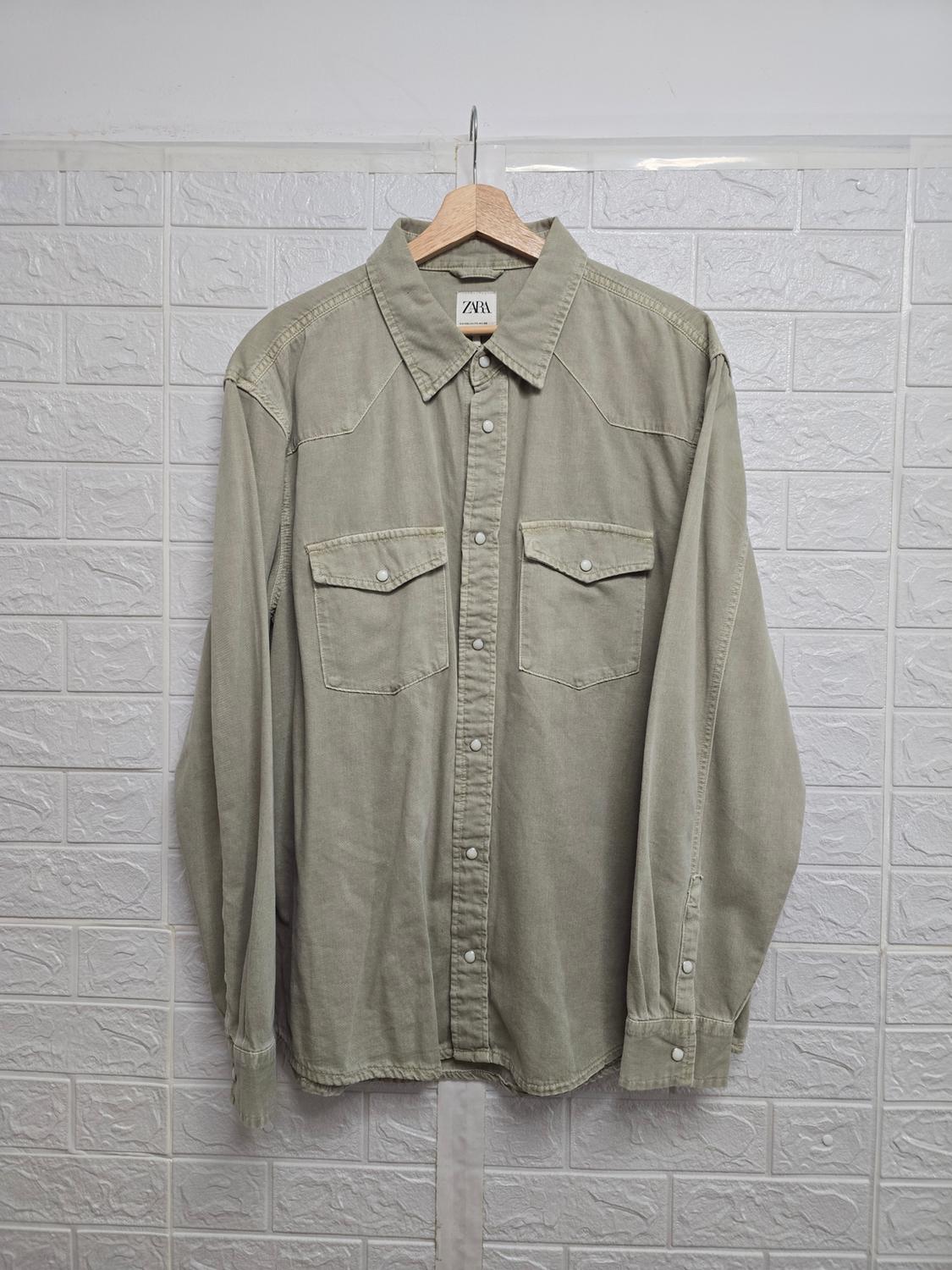 Olive green, long-sleeved shirt with front pockets and buttons, hanging on a wooden hanger. Visible label: "ZARA".