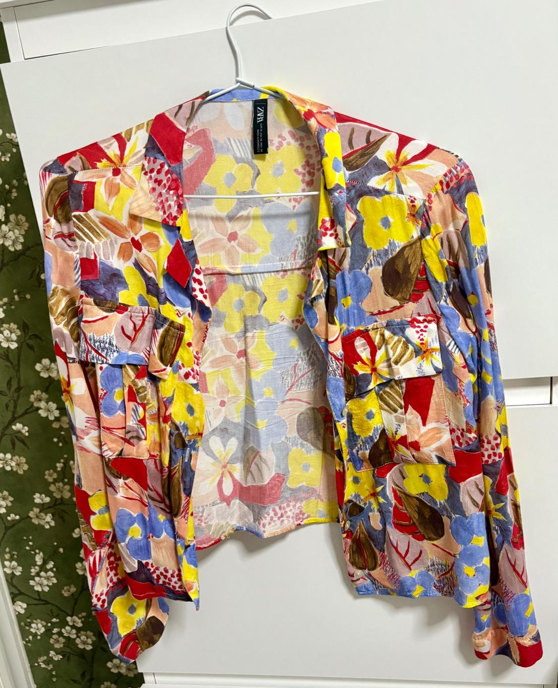 Hanging shirt with a floral print in yellow, red, and blue tones. White and green background with small flowers. Label visible.