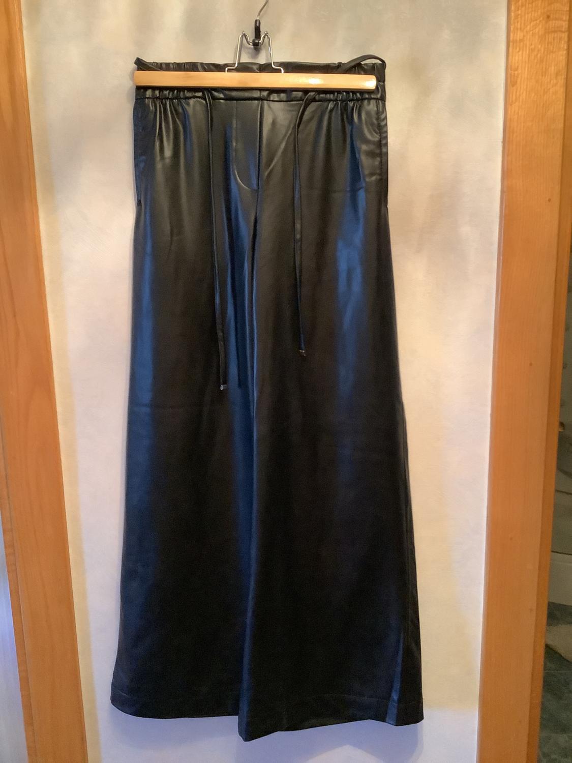 Black long trousers hung on a wooden hanger, with drawstrings at the waist, against a light background and wooden frame.