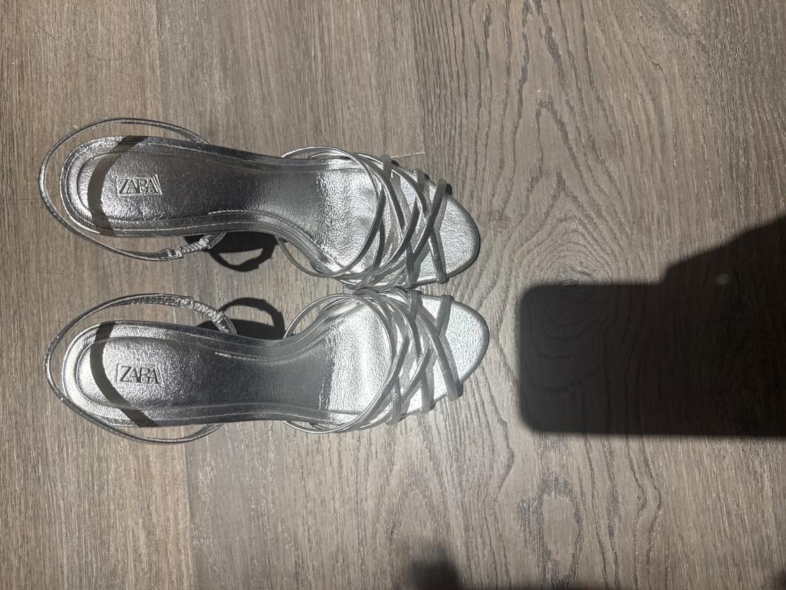 Silver high-heeled sandals with crossed straps on a wooden floor. Visible text: "ZARA". Shadow of a person in the image.