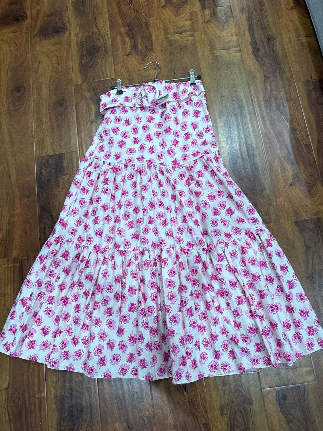 White tiered maxi skirt with a pink floral print and belt. It is hanging on a hanger above a wooden floor.