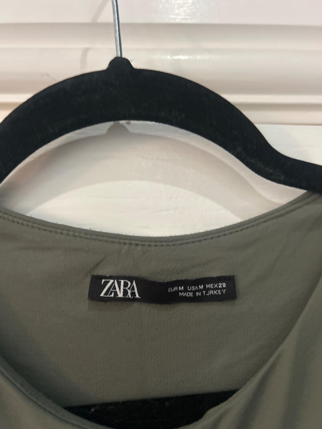 Label of a green garment on a black hanger. Visible text: "ZARA EUR M USA M MEX 28 MADE IN TURKEY". White background.
