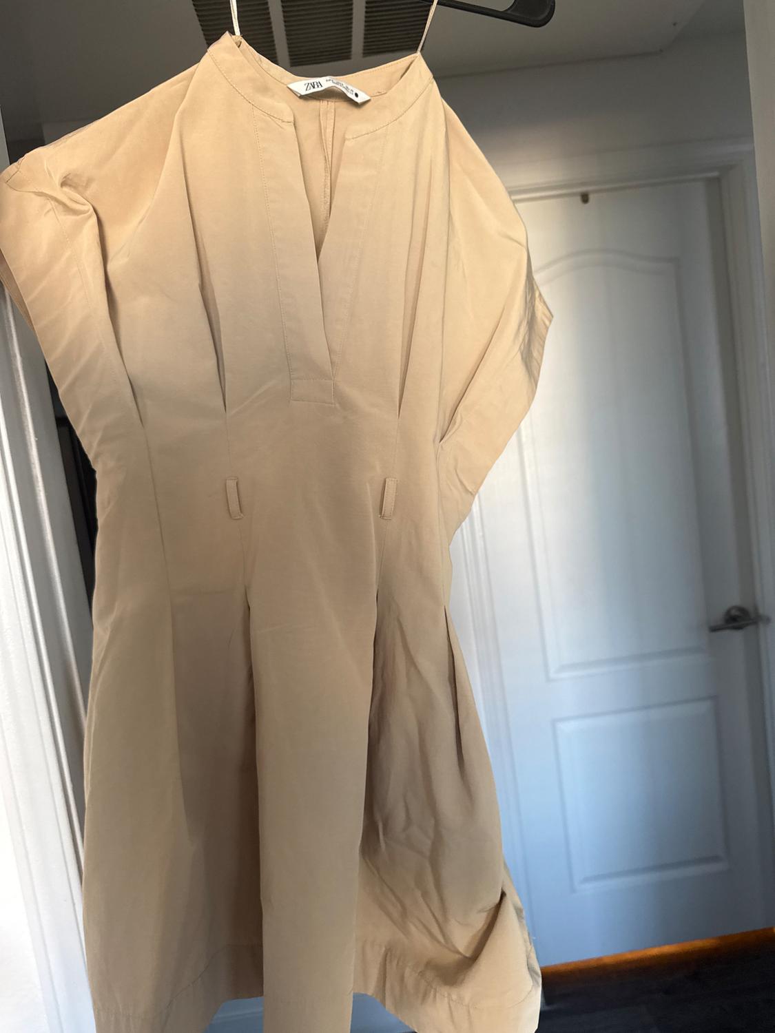 Beige sleeveless dress with a V-neck and pleats at the waist, hanging indoors against a white door. Text ZARA.