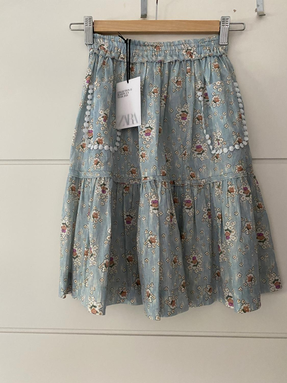 Blue floral print skirt with pom-pom pockets hanging on a wall. Text: COLLECTION 15 SEASON S/S YEAR 2024 ZARA.