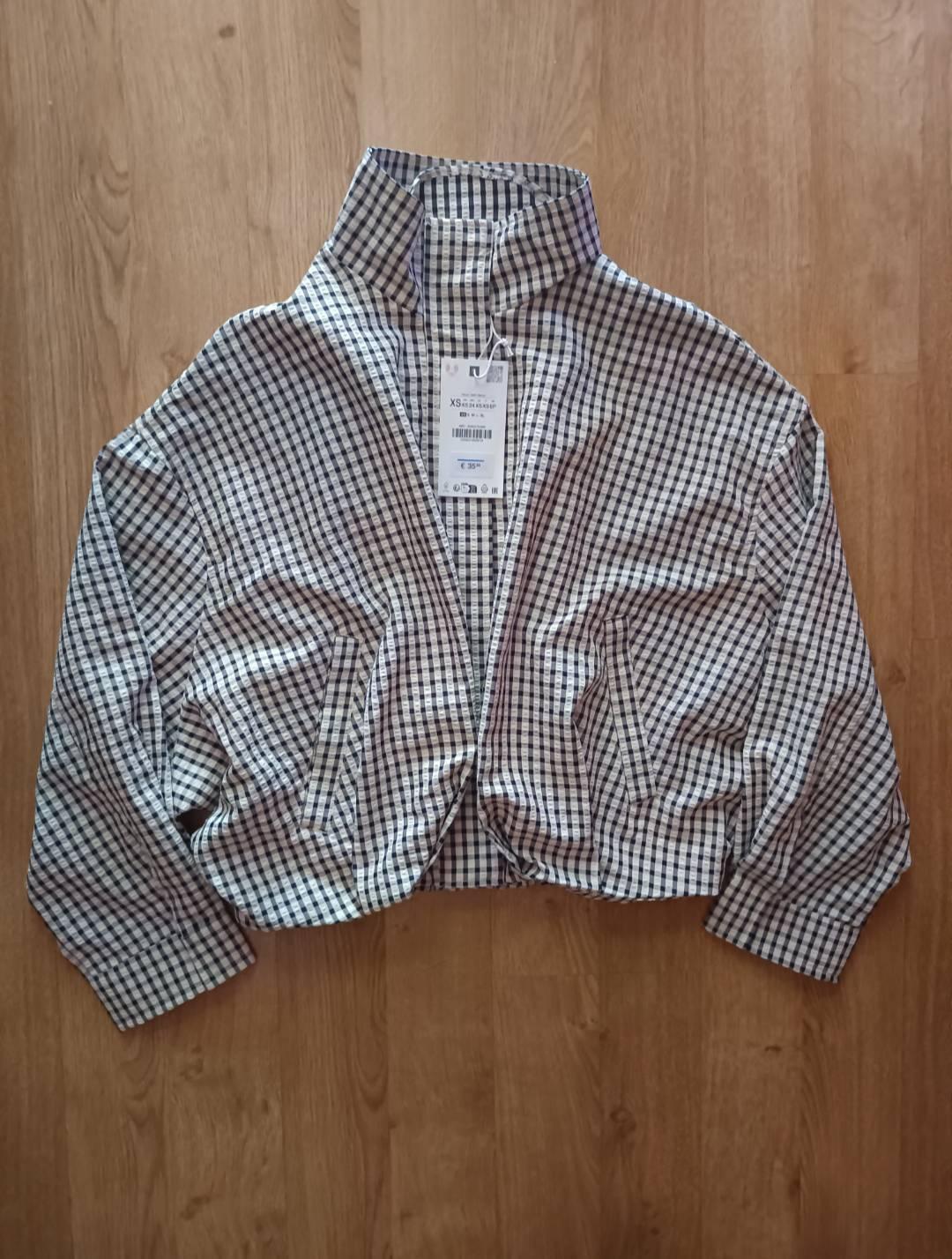 Black-and-white checked jacket with a high collar on a wooden floor. Visible label bearing the text “XS” and “€ 35”.