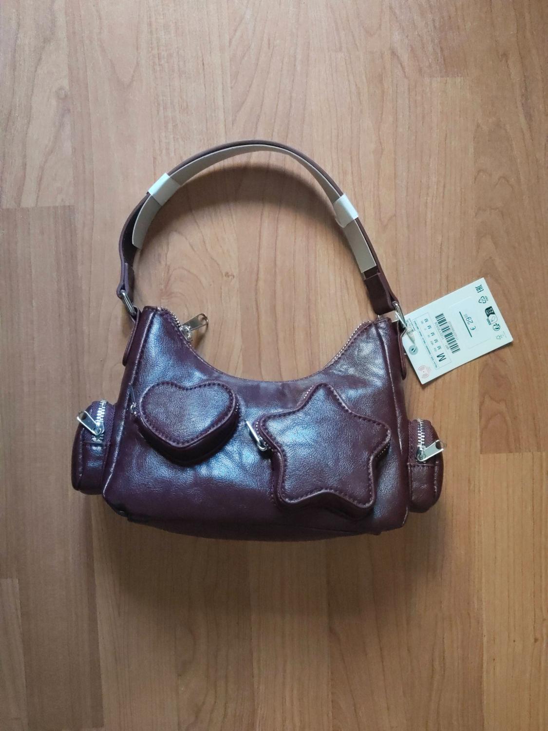 Maroon shoulder bag on wood with heart- and star-shaped front pockets. Label with text M M M M M M €29.95.