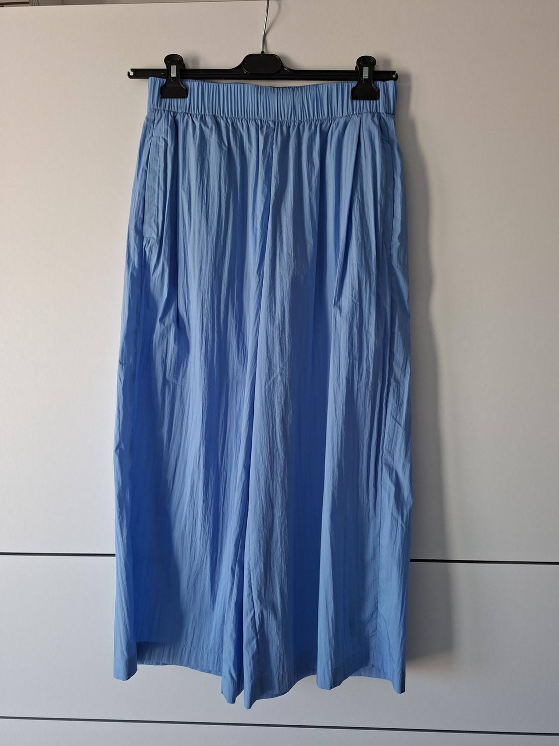 Light blue wide-leg trousers with an elasticated waist and crinkled texture, hanging on a black hanger in front of a white wardrobe.