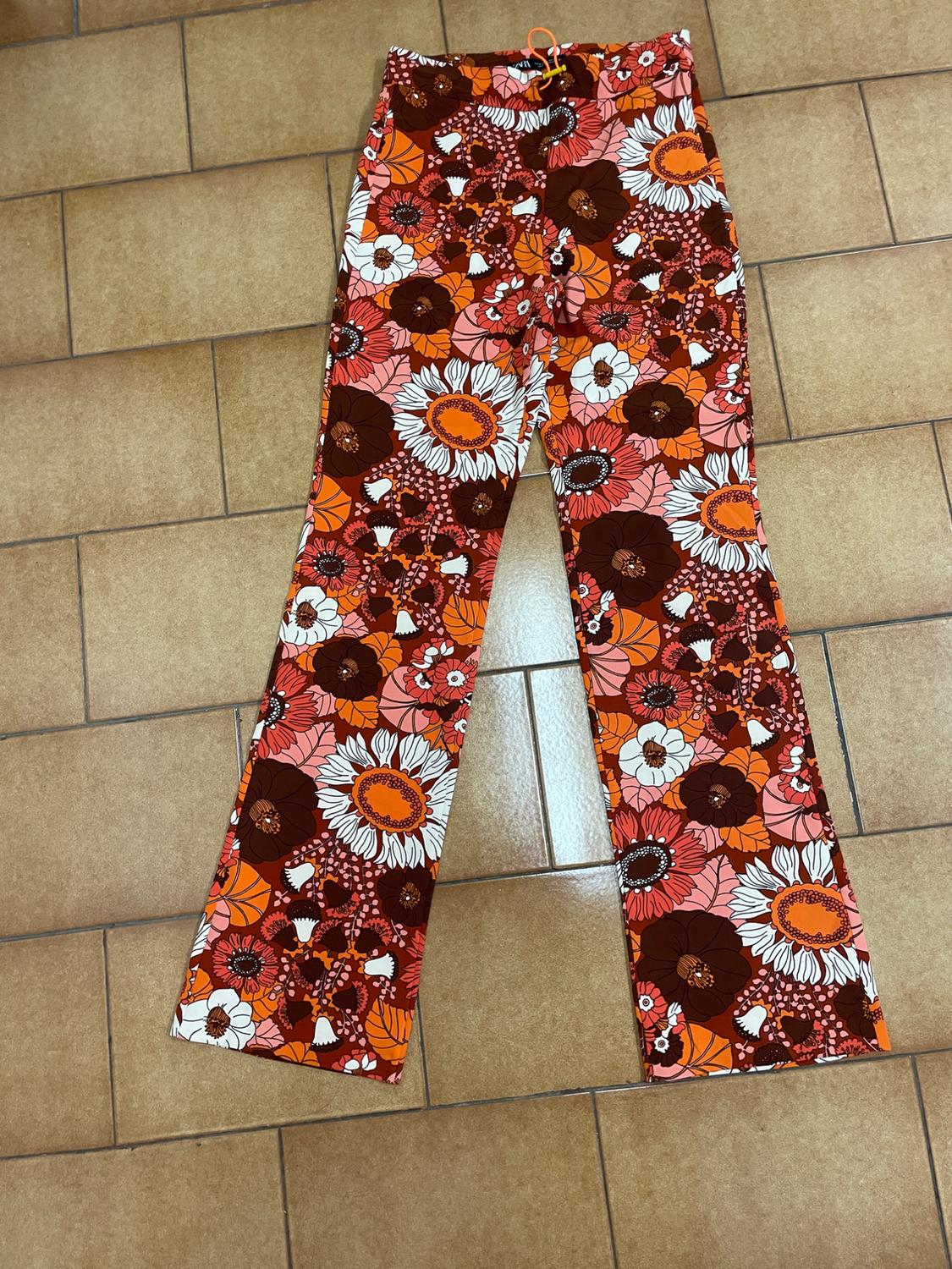 Flared trousers with a floral print in shades of brown, orange, and white on a light brown tiled floor.