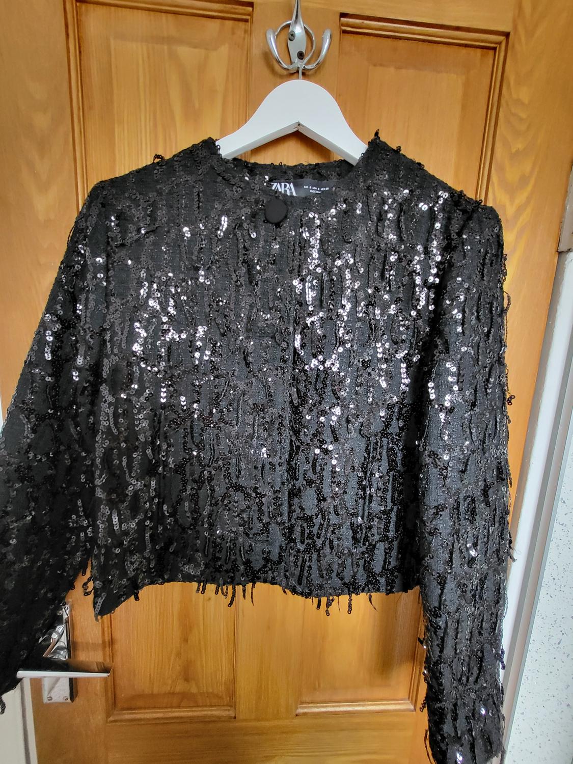 Black sequinned jacket hanging on a white hanger on a wooden door. Inside label with the text ZARA.