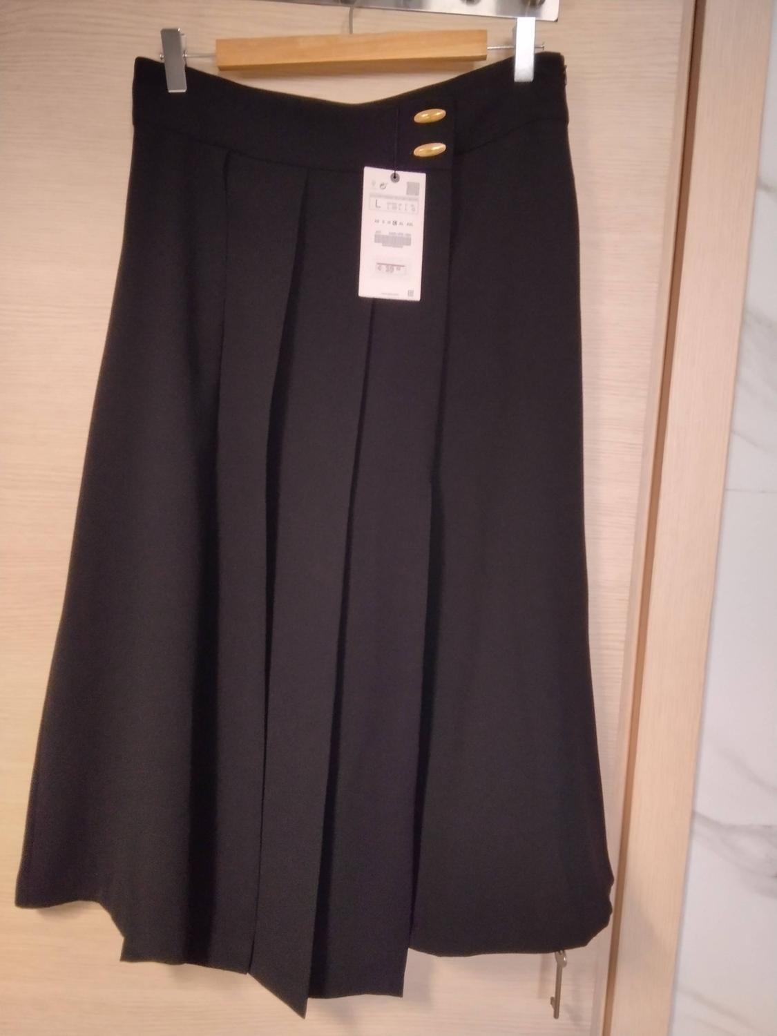 Black pleated skirt with gold buttons at the waist, hanging on a wooden hanger with clips in front of a light-coloured door.