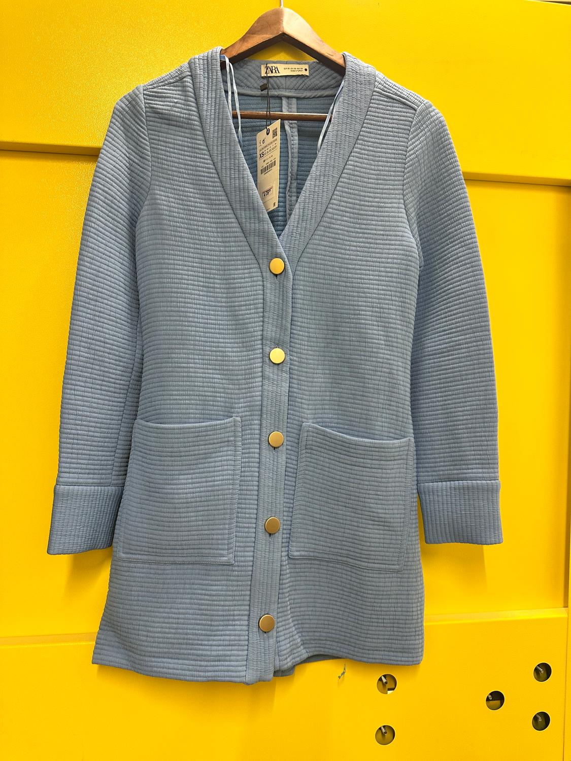 Light blue coat with gold buttons and large pockets, hanging on a hanger against a bright yellow background.