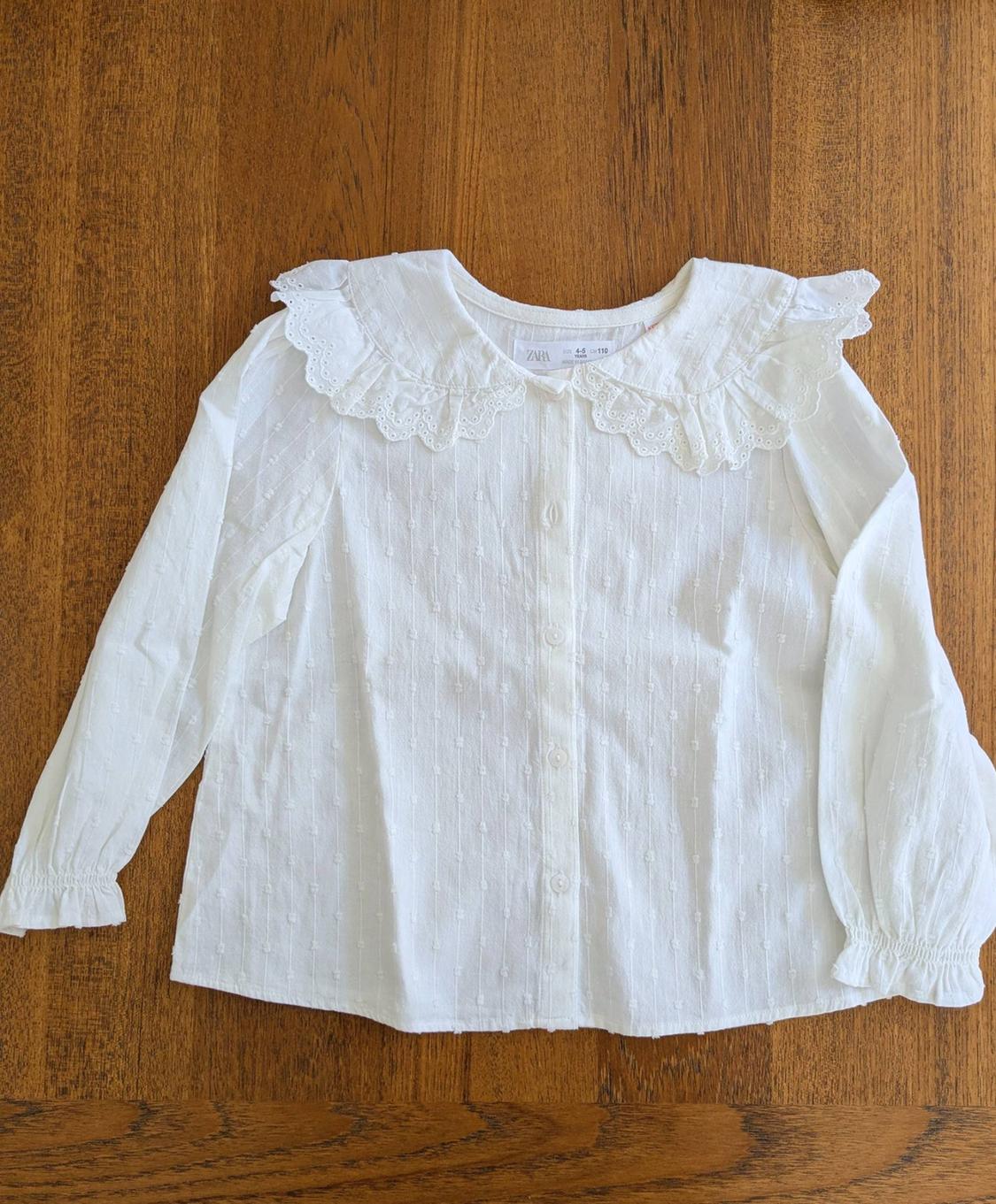 White long-sleeved blouse with a ruffled collar and embroidered details, on a wooden surface. Visible tag: ZARA.