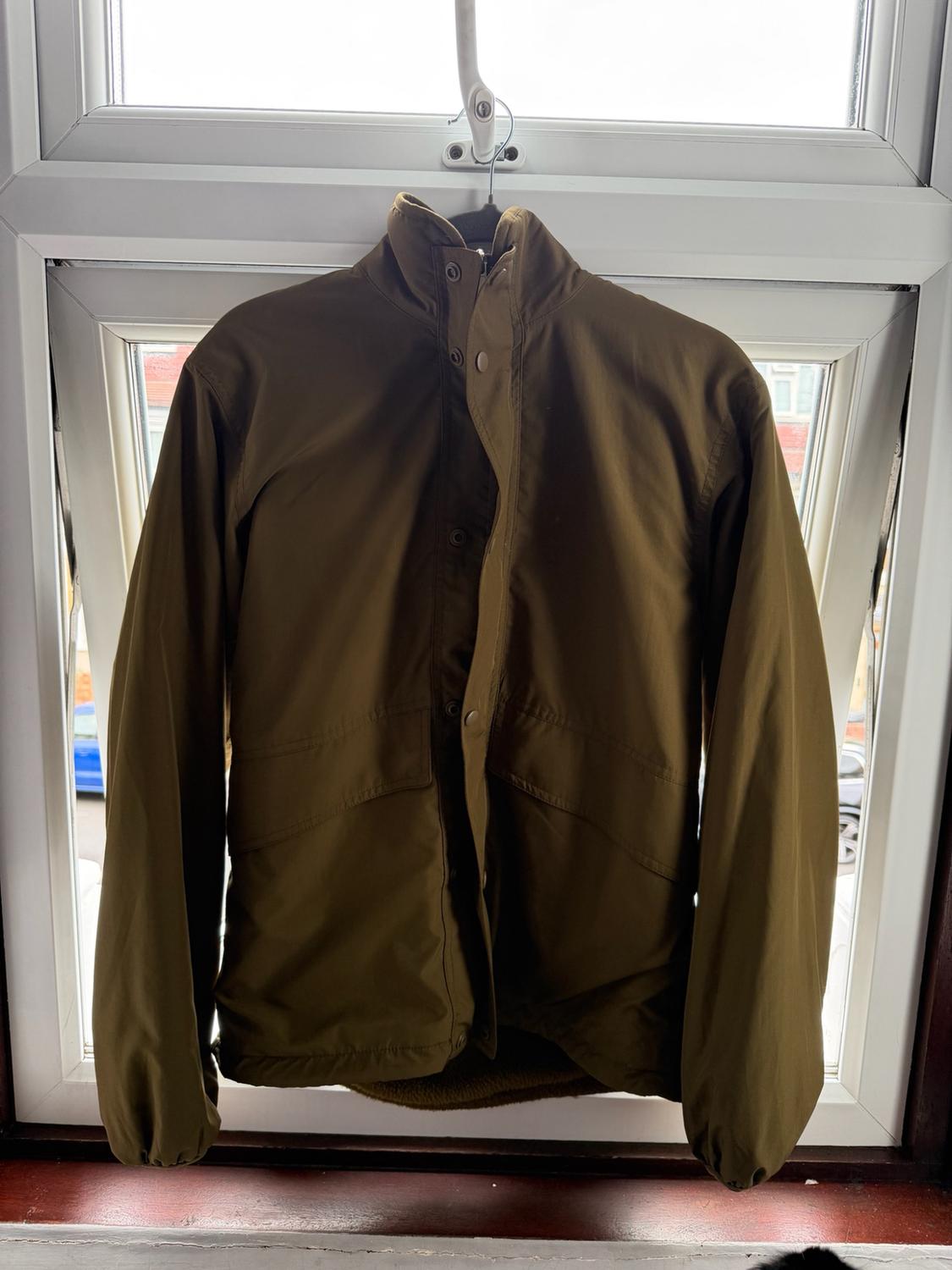 Olive green casual jacket with a high collar and front buttons, hanging on a hanger in front of a white window.