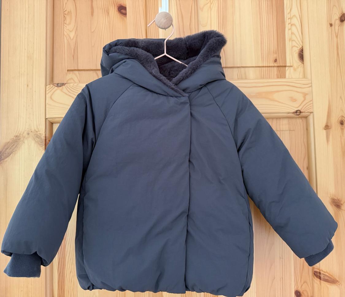 Dark blue quilted coat with a hood and elasticated cuffs, hanging on a hanger over a light wood door.