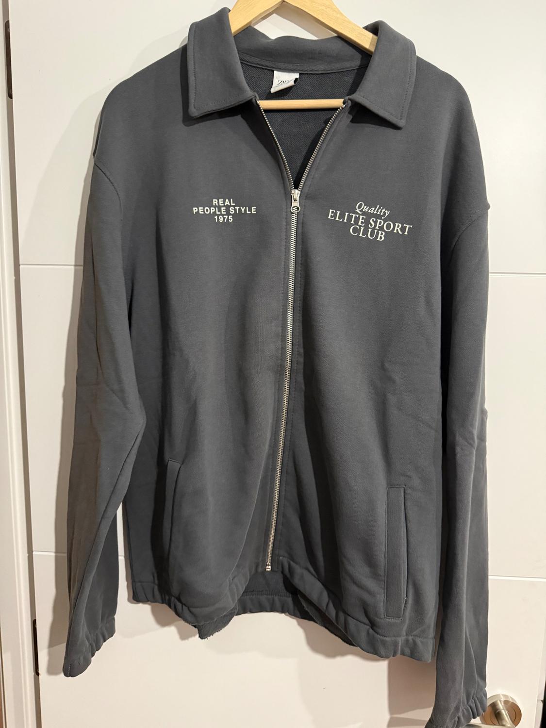 Grey zip-up jacket hanging on a hanger. Text: "REAL PEOPLE STYLE 1975" and "Quality ELITE SPORT CLUB" on the chest.