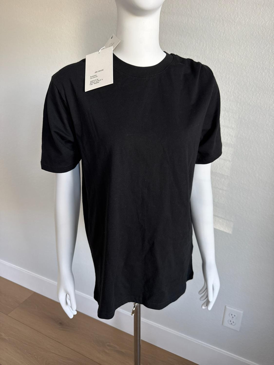White mannequin wearing a short-sleeved black t-shirt and a tag with the text ZARA ORIGINS. Light wall background and wooden floor.