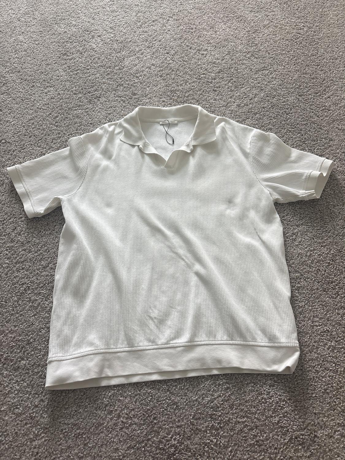 Short-sleeved white collared shirt, placed on a gray rug. Fabric with subtle texture and cuffed sleeves.
