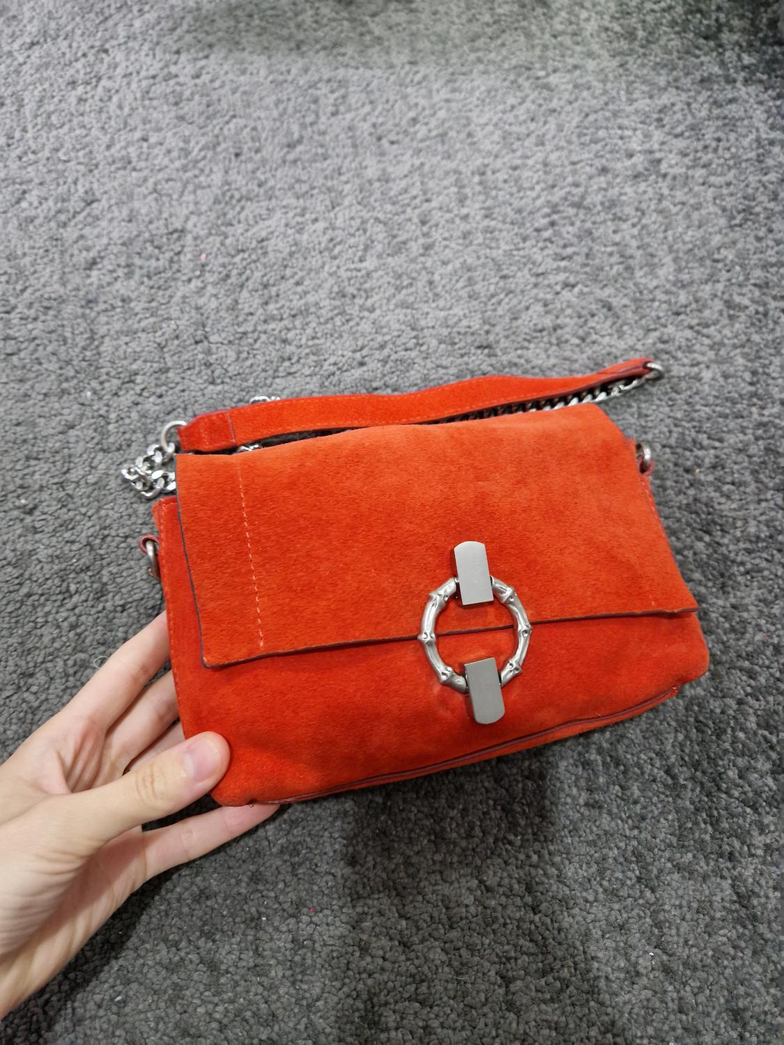 Hand holding an orange suede handbag with a silver ring clasp and chain strap over a grey carpet.