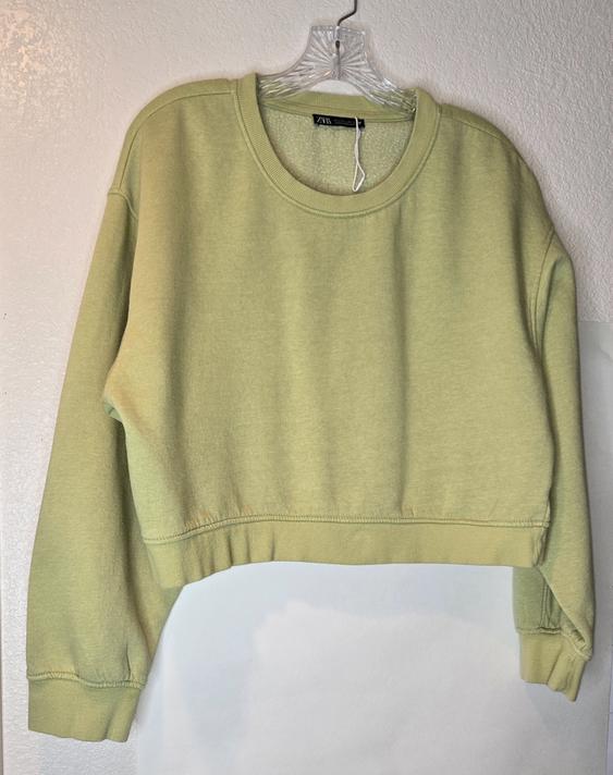 Light green cropped sweatshirt hanging on a clear hanger against a white wall. Visible tag on the collar.