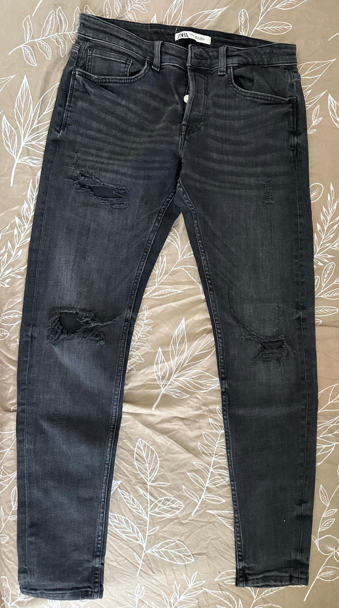 Distressed black jeans with rips and a ZARA label, on a beige fabric with a white leaf print.